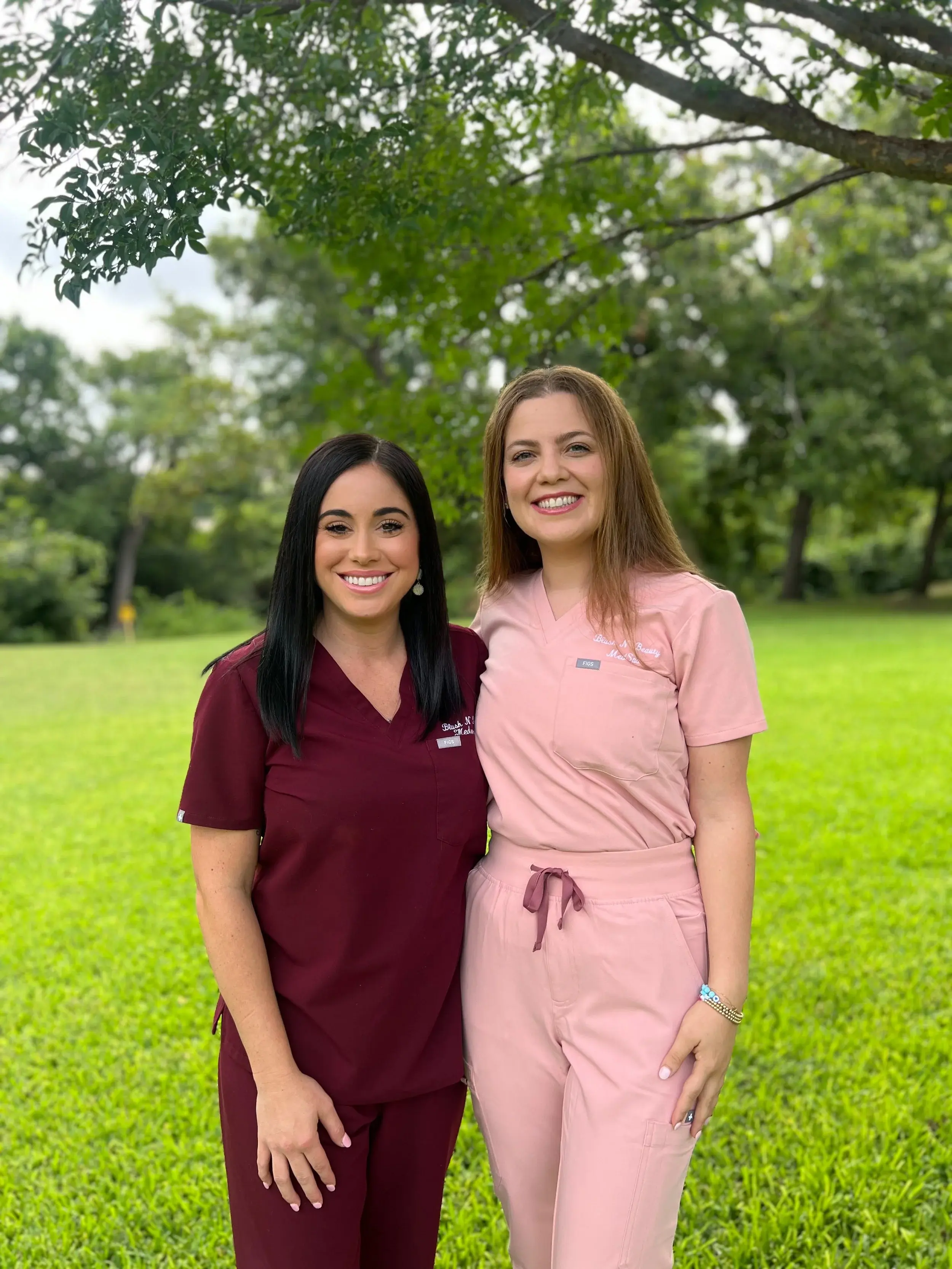 Two women in medical scrubs standing outdoors on a grassy area with trees in the background, smiling at the camera.